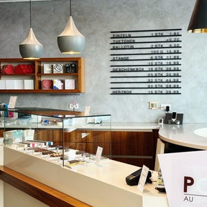 May include: A retail chocolate shop interior featuring a glass display counter filled with assorted chocolates, a wooden wall shelf with boxed items, and a black wall-mounted menu board listing items like Einzeln, Tuetchen, Ballotin, Stange, and Herzbox.
