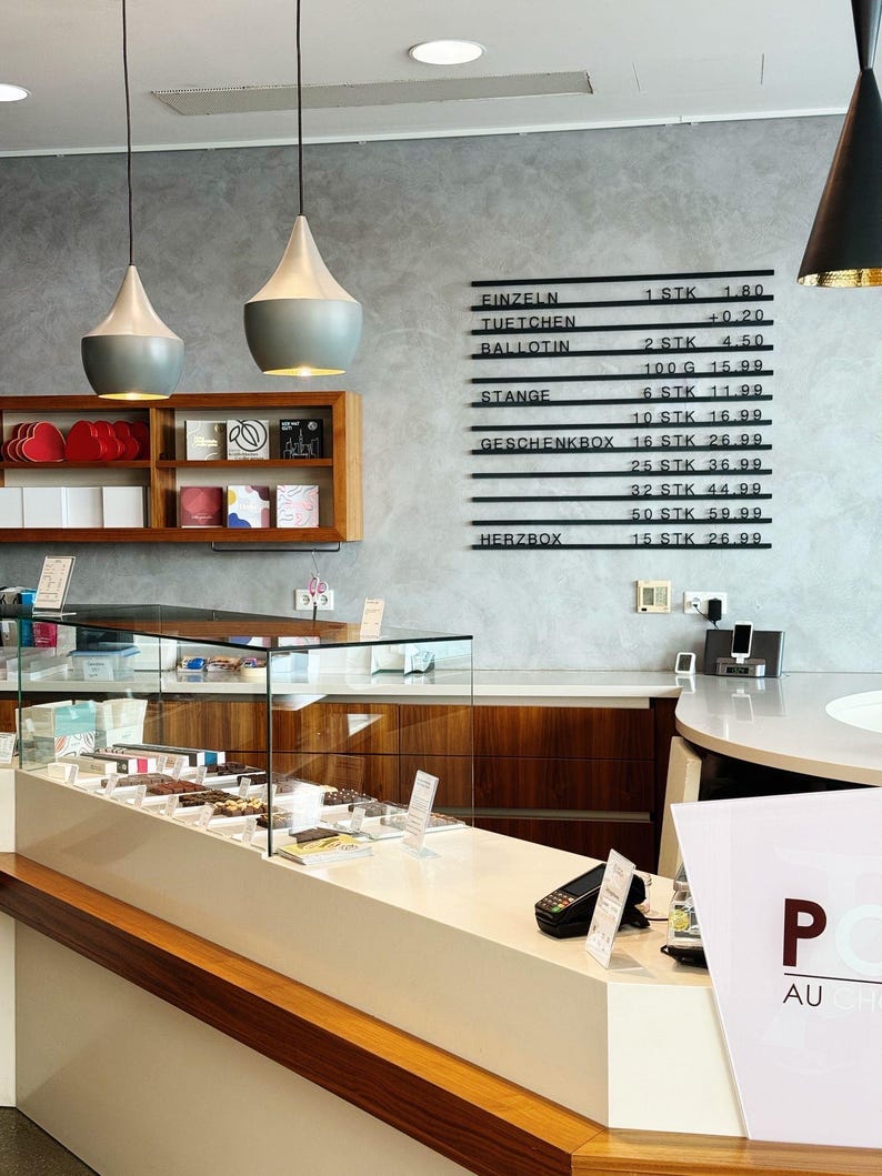 May include: A retail chocolate shop interior featuring a glass display counter filled with assorted chocolates, a wooden wall shelf with boxed items, and a black wall-mounted menu board listing items like Einzeln, Tuetchen, Ballotin, Stange, and Herzbox.
