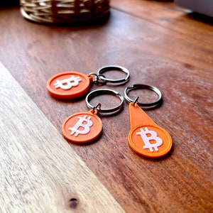 Pode incluir: Três chaveiros laranja com um símbolo Bitcoin branco. Os chaveiros estão sobre uma superfície de madeira.
