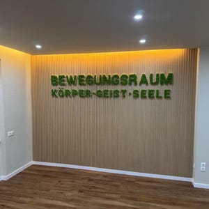 Pu&ograve; includere: Vista interna di una stanza con pavimenti e pareti in legno. La parete presenta pannelli di legno verticali e il testo "BEWEGUNGSRAUM K&Ouml;RPER&bull;GEIST&bull;SEELE" in lettere verdi, simili a muschio. L'illuminazione a incasso illumina lo spazio.