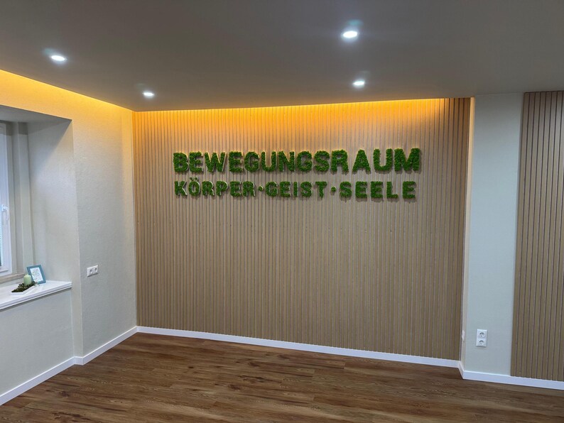 Pu&ograve; includere: Vista interna di una stanza con pavimenti e pareti in legno. La parete presenta pannelli di legno verticali e il testo "BEWEGUNGSRAUM K&Ouml;RPER&bull;GEIST&bull;SEELE" in lettere verdi, simili a muschio. L'illuminazione a incasso illumina lo spazio.