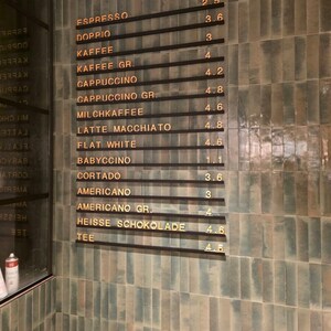 May include: A wall-mounted menu board featuring horizontal black rails with gold lettering listing coffee drinks including Espresso, Doppio, Kaffee, Cappuccino, Milchkaffee, Latte Macchiato, Flat White, Babyccino, Cortado, Americano, Heisse Schokolade, and Tee.