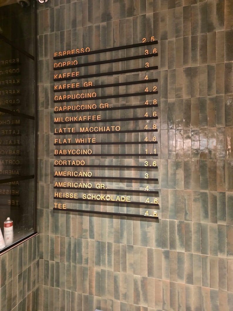 May include: A wall-mounted menu board featuring horizontal black rails with gold lettering listing coffee drinks including Espresso, Doppio, Kaffee, Cappuccino, Milchkaffee, Latte Macchiato, Flat White, Babyccino, Cortado, Americano, Heisse Schokolade, and Tee.