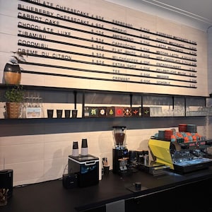 May include: A cafe counter featuring a large wall-mounted menu board with black horizontal slats displaying various food and drink items with prices in euros. Below, a black shelf holds plants and cups, while an espresso machine sits on the counter.