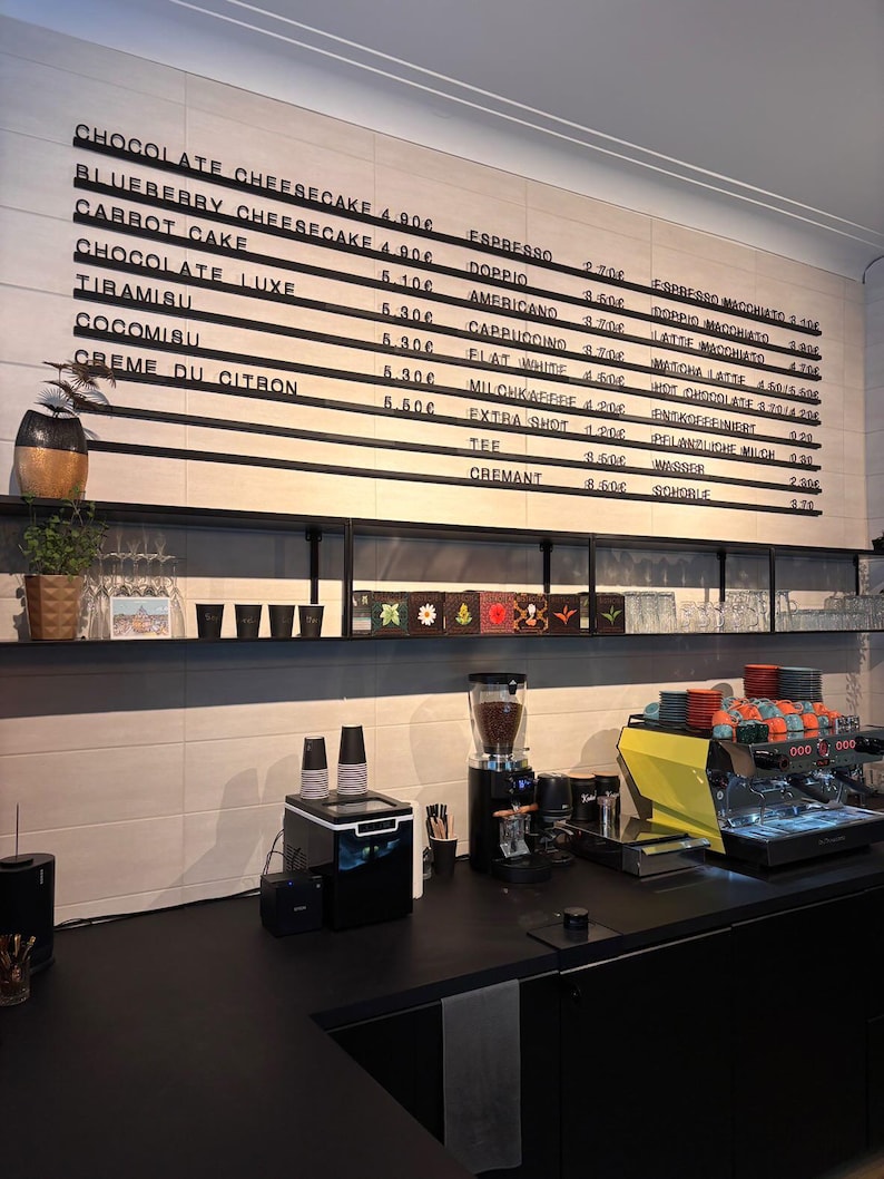 May include: A cafe counter featuring a large wall-mounted menu board with black horizontal slats displaying various food and drink items with prices in euros. Below, a black shelf holds plants and cups, while an espresso machine sits on the counter.