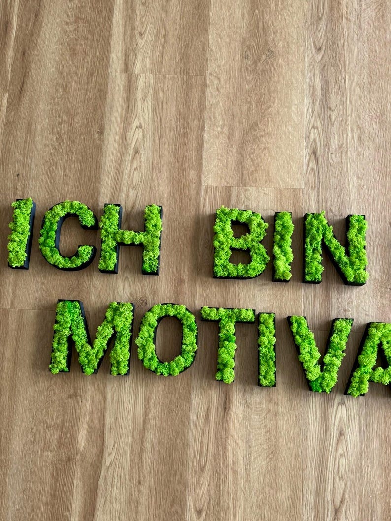 Pu&ograve; includere: Lettere di muschio verde che compongono la scritta "ICH BIN MOTIVAT" su una superficie di legno. Le lettere sono delineate in nero, con il muschio che riempie l'interno di ogni lettera. L'estetica generale &egrave; naturale e decorativa.