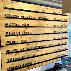 May include: A light-colored wooden menu board with horizontal black rails holding black plastic letters. The text lists coffee varieties including Crema Monaco Arabica, Extra Bar, Finest India, Peru Bio, Schoene Muenchnerin Bio, Premium Decaf, and Probierpaket.