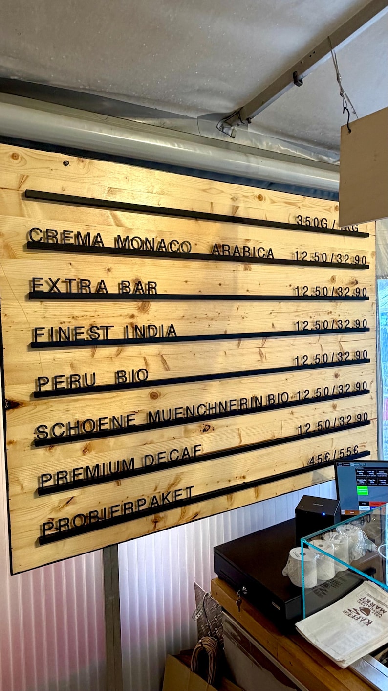 May include: A light-colored wooden menu board with horizontal black rails holding black plastic letters. The text lists coffee varieties including Crema Monaco Arabica, Extra Bar, Finest India, Peru Bio, Schoene Muenchnerin Bio, Premium Decaf, and Probierpaket.