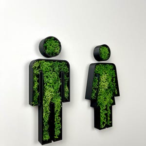 May include: Black and green wall mounted restroom signs with moss inside the cutouts. The signs are shaped like stick figures.
