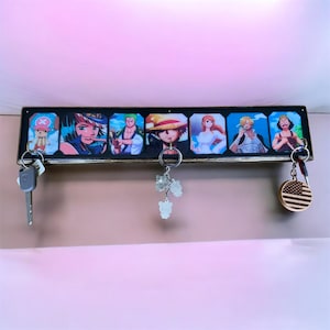 Puede incluir: Un soporte de pared rectangular para llaves que presenta una serie de coloridas ilustraciones de personajes de anime en paneles individuales. El estante de madera incluye tres ganchos metálicos, con una llave plateada, un dije de oso.