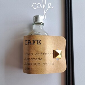 Könnte beinhalten: Ein braunes Papieretikett mit dem Wort "CAFE" in goldenen Buchstaben, mit dem Text "reed diffuser", "handmade", "SANAHOM brand" und "30ml" darauf gedruckt. Eine klare Glasflasche mit silbernem Deckel und schwarzen Schilfrohrstäben sind sichtbar.