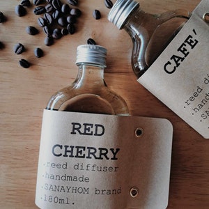 Könnte beinhalten: Zwei Glasflaschen mit braunen Papierretiketten. Die Flasche links ist mit "RED CHERRY reed diffuser handmade SANAYHOM brand 180ml." beschriftet. Die Flasche rechts ist mit "CAFE' reed di hand SA" beschriftet. Beide Flaschen stehen auf einer Holzoberfläche mit verstreuten Kaffeebohnen.