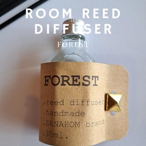 May include: A clear glass room reed diffuser bottle with a silver cap and a brown label. The label reads "FOREST" and "SANAYHOM brand". The bottle is accompanied by black reed sticks and a gold stud.
