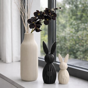Könnte beinhalten: Eine cremefarbene Vase mit vertikalen Rippen enthält getrocknete Zweige und dunkelviolette Orchideen. Zwei dekorative Hasenfiguren, eine schwarze und eine cremefarbene, mit Herzdetails, sitzen auf einer Fensterbank. Die Szene spielt sich vor einem Fenster mit Blick ins Grüne ab.