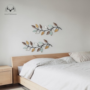 Pode incluir: Duas peças de arte de parede de galhos de folhas de metal. Cada galho tem um caule preto com folhas multicoloridas em tons de marrom, dourado, branco e azul. A arte de parede é exibida acima de uma estrutura de cama de madeira com um edredom e travesseiro brancos.
