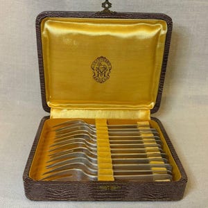 May include: A set of silver forks in a brown, textured case with a yellow velvet interior. The case is open, revealing the forks neatly arranged. The case has a gold clasp and a decorative emblem.