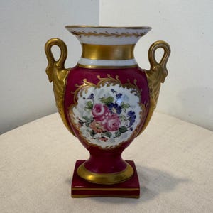 Puede incluir: Un jarrón decorativo con base cuadrada, con cuerpo rojo oscuro y detalles dorados. El jarrón tiene dos asas ornamentadas y un diseño floral en el centro, con flores rosas y azules. El borde superior es blanco con detalles dorados.