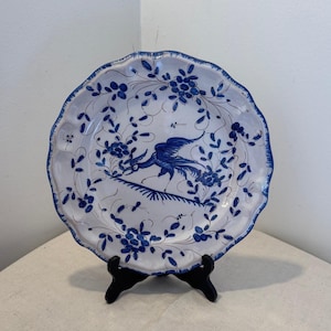 Può includere: Un piatto rotondo in ceramica con bordo smerlato, caratterizzato da una base bianca decorata con motivi floreali blu dipinti a mano e un'illustrazione centrale di un uccello. Il bordo è accentuato da una bordatura blu e il piatto poggia su un supporto nero.