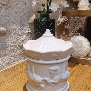 Op de afbeelding: Witte keramische kaarshouder in de vorm van een draaimolen met paarden. De kaarshouder heeft een wit deksel met een decoratief bovendeel.