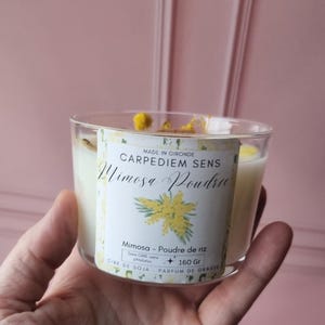 May include: A white candle in a glass jar with a label that reads "CARPEDIEM SENS Mimosa Poudree". The label also includes the text "Mimosa - Poudre de riz", "CIRE DE SOJA", "PARFUM DE GRASSE", and "160 Gr".