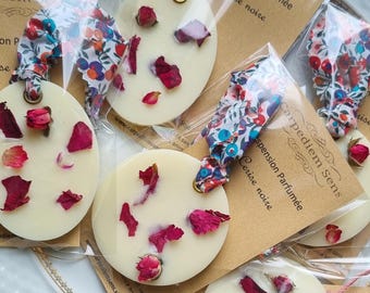 Può includere: Quattro sciogli-profumo rotondi di cera bianca con petali e boccioli di rosa secchi. Ogni sciogli-profumo è confezionato in un sacchetto di plastica trasparente con un'etichetta di carta marrone che recita "Carpe diem Sens Suspension Parfumée Cerise noire" e un indirizzo di sito web. Gli sciogli-profumo sono su un piatto bianco con un bordo dorato.