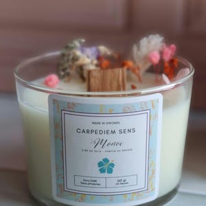 May include: A white soy wax candle with a blue and white label. The label reads "CARPEDIEM SENS Monoi CIPE DE SOJA PARFUM DE GRASSE Sans CMR Sans phtalates 160 gr 45 Heures". The candle is decorated with dried flowers.