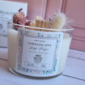 Pu&ograve; includere: Una candela bianca di cera di soia in un barattolo di vetro trasparente con un'etichetta decorativa. L'etichetta dice "Carpediem Sens Linge Propre" e "Cire de Soja - Parfum de Grasse". La candela &egrave; decorata con fiori secchi e un bastoncino di legno.