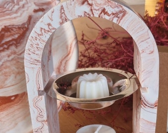 Può includere: Un portacandele decorativo con un design bianco e rosa marmorizzato. Il supporto presenta una parte superiore ad arco e un piatto d'argento che contiene una candela bianca. Sotto c'è una candela da tè più piccola. Sono visibili rami rossi secchi decorativi.