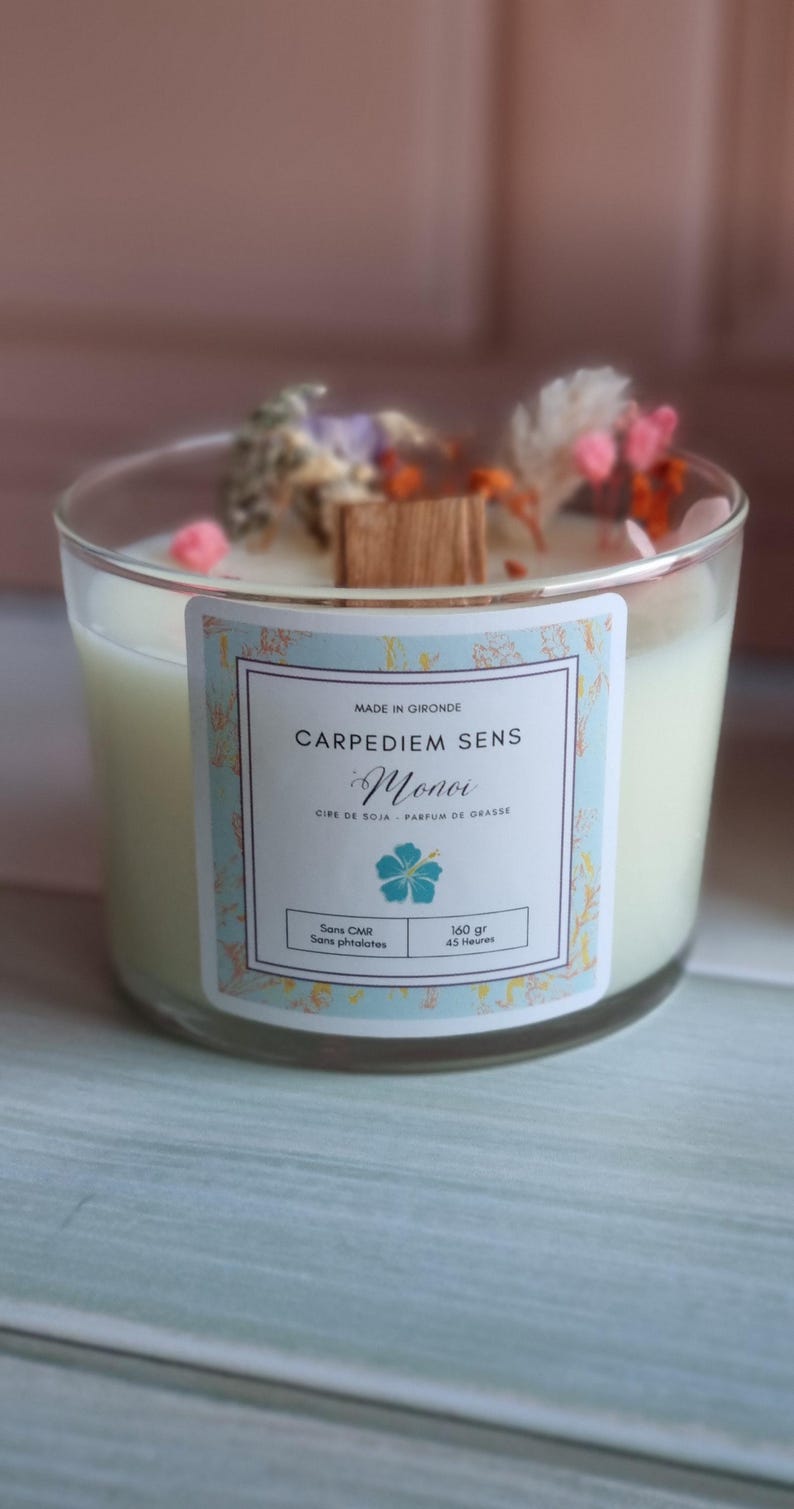 Pu&ograve; includere: Una candela in vetro trasparente con una base di cera di soia bianca e un'etichetta che recita "CARPEDIEM SENS Monoi CIPE DE SOJA - PARFUM DE GRASSE Sans CMR Sans phtalates 160 gr 45 Heures". La candela &egrave; decorata con fiori secchi e uno stoppino di legno.