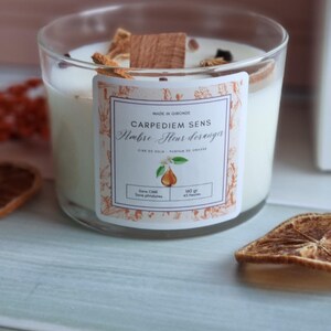 Pu&ograve; includere: Una candela bianca di cera di soia in un barattolo di vetro trasparente con un'etichetta che recita "Carpediem Sens,  Ambre Fleur d'oranger, Cire de Soja, Parfum de Grasse, Sans CMR, Sans phtalates, 140 gr, 45 heures". La candela &egrave; circondata da fette di arancia essiccate e un bastoncino di legno.