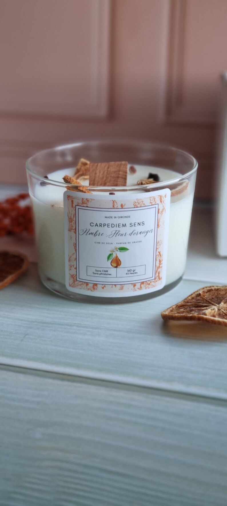 Pu&ograve; includere: Una candela bianca di cera di soia in un barattolo di vetro trasparente con un'etichetta che recita "Carpediem Sens,  Ambre Fleur d'oranger, Cire de Soja, Parfum de Grasse, Sans CMR, Sans phtalates, 140 gr, 45 heures". La candela &egrave; circondata da fette di arancia essiccate e un bastoncino di legno.