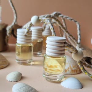 Può includere: Tre piccole bottiglie di vetro con tappi di legno e uno spago beige attaccato. Le bottiglie sono riempite con un liquido giallo chiaro. Le bottiglie sono circondate da conchiglie bianche su una superficie beige.