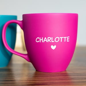 Könnte beinhalten: Ein leuchtend pinkfarbener Keramikbecher mit matter Oberfläche, auf dem der Name CHARLOTTE in weißen Großbuchstaben über einem kleinen weißen Herzsymbol aufgedruckt ist. Der Becher hat einen geschwungenen Henkel und steht zwischen einem türkisen und einem dunkelblauen Becher.