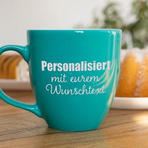 Könnte beinhalten: Eine türkisfarbene Keramiktasse mit gebogenem Henkel. Die Tasse trägt den weißen Text "Personalisiert mit eurem Wunschtext". Ein Kuchen und Gebäck sind im Hintergrund zu sehen.