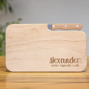 Könnte beinhalten: Ein helles Holzschneidebrett mit einem kleinen Messer. Das Brett hat abgerundete Ecken und den Namen "Alexander" und den Satz "bester Papa der Welt!" eingraviert. Das Messer hat einen Holzgriff und eine silberne Klinge.