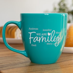 Könnte beinhalten: Ein türkisfarbener Keramikbecher mit dem weißen Aufdruck WE ARE Family, einem kleinen Herzsymbol und den Namen Andreas, Jennifer, Max, Emil und Mary, die um das zentrale Wort angeordnet sind. Der Becher hat einen geschwungenen Henkel und eine glatte Oberfläche.