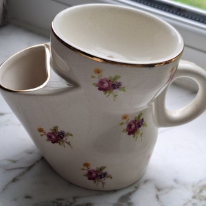 Op de afbeelding: Crèmekleurige scheerkop met een ingebouwde zeepbakje. De kop heeft een gouden rand en is versierd met kleine bloemboeketten in paars, roze en geel. Het heeft een gebogen handvat en staat op een wit oppervlak.