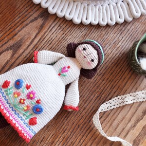 May include: A handmade crochet doll in a white dress with colourful floral embroidery. The doll has brown hair, a mint green headband, and red sleeve accents. The doll is lying on a wooden surface.