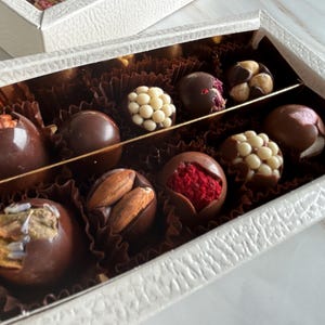 Puede incluir: Una caja blanca texturizada llena de trufas de chocolate variadas. Las trufas son de color marr&oacute;n oscuro y est&aacute;n decoradas con nueces, perlas de chocolate blanco y fruta roja. La caja est&aacute; dividida en secciones con un divisor dorado.