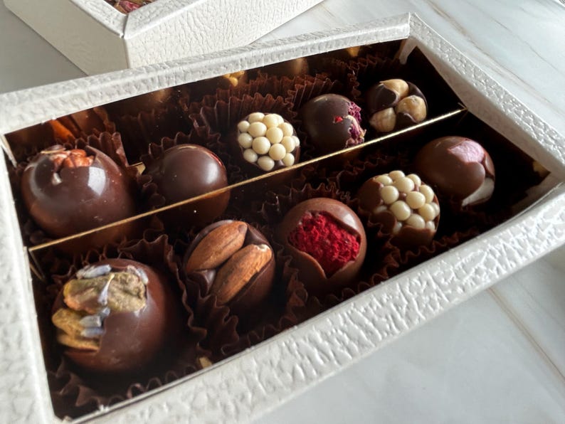 Puede incluir: Una caja blanca texturizada llena de trufas de chocolate variadas. Las trufas son de color marr&oacute;n oscuro y est&aacute;n decoradas con nueces, perlas de chocolate blanco y fruta roja. La caja est&aacute; dividida en secciones con un divisor dorado.