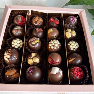 Puede incluir: Una caja rosa llena de bombones variados. Cada bomb&oacute;n es de chocolate negro y est&aacute; decorado con frutos secos, frutas deshidratadas y otros ingredientes. Los bombones est&aacute;n dispuestos en copas de papel individuales dentro de la caja.