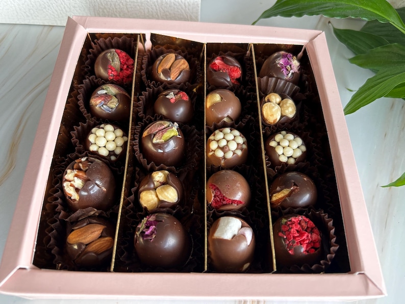 Puede incluir: Una caja rosa llena de bombones variados. Cada bomb&oacute;n es de chocolate negro y est&aacute; decorado con frutos secos, frutas deshidratadas y otros ingredientes. Los bombones est&aacute;n dispuestos en copas de papel individuales dentro de la caja.