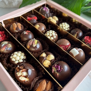 Könnte beinhalten: Eine rosa Schachtel gefüllt mit verschiedenen Schokoladentrüffeln. Die Trüffel sind aus dunkler Schokolade und mit Nüssen, Samen und Trockenfrüchten verziert. Die Schachtel ist in Abschnitte unterteilt, die jeweils eine andere Trüffelart enthalten.