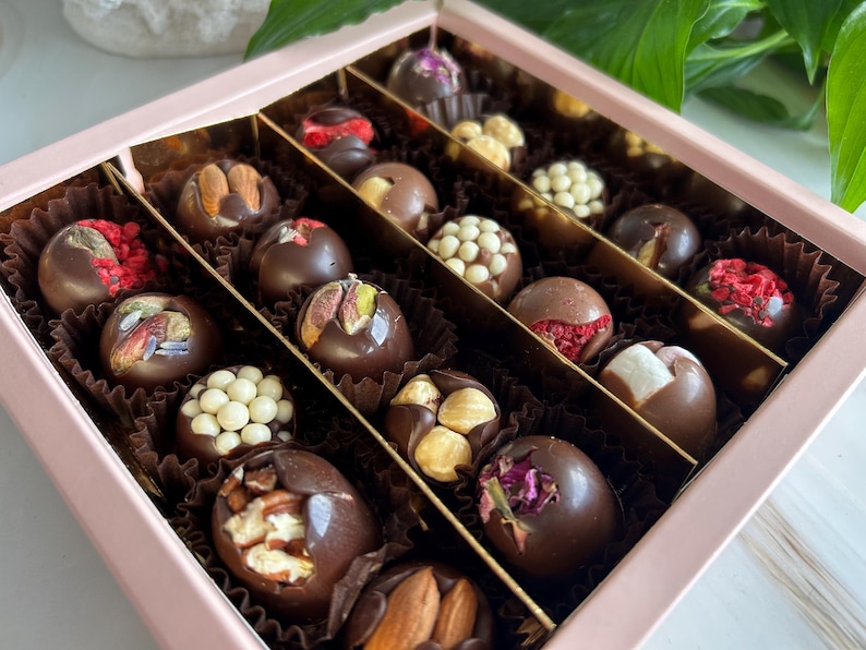 Puede incluir: Una caja rosa llena de trufas de chocolate surtidas. Las trufas son de chocolate negro y est&aacute;n decoradas con frutos secos, semillas y frutas deshidratadas. La caja est&aacute; dividida en secciones, cada una con un tipo diferente de trufa.