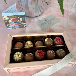 Puede incluir: Una caja de bombones de chocolate surtidos con varios ingredientes, incluyendo nueces y bayas secas. La caja es de color rosa claro con una cinta blanca que dice "Petal & Cocoa". Se ve una peque&ntilde;a tarjeta con una manzana y libros.