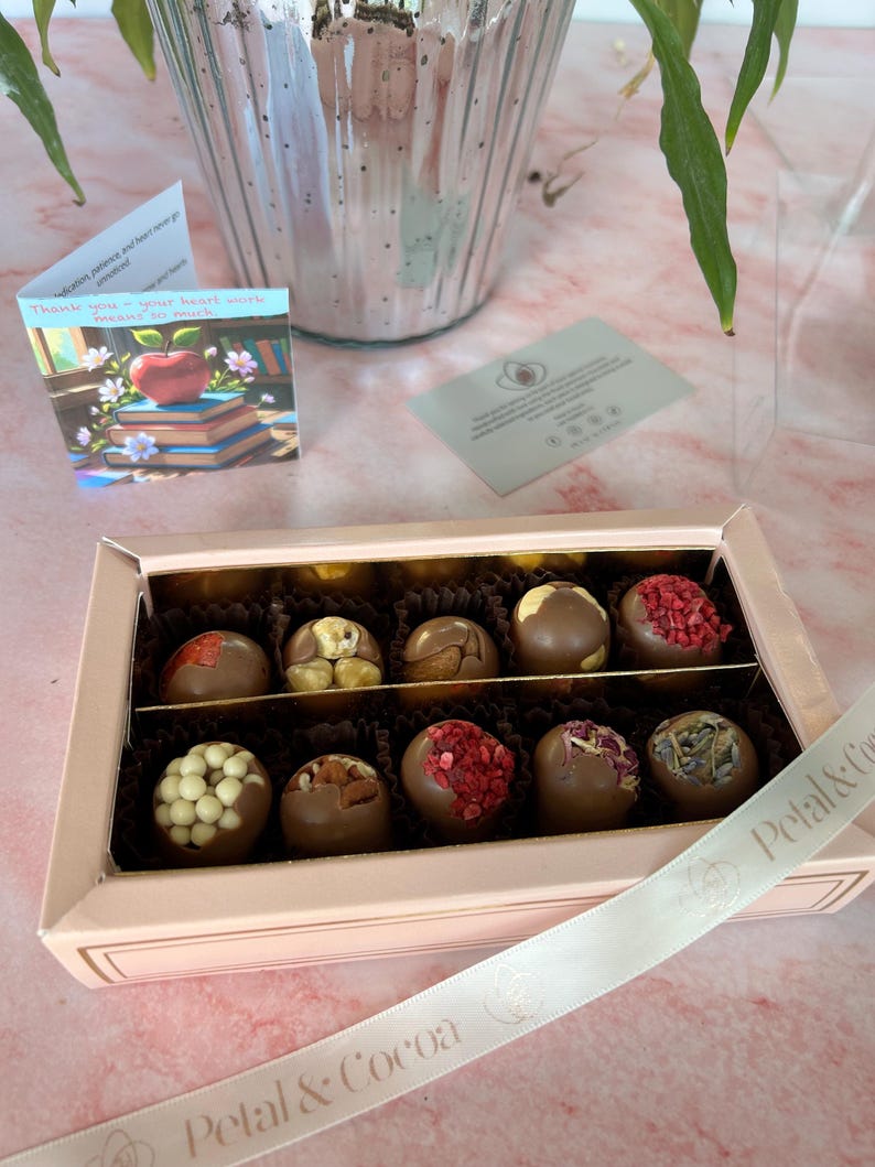 Puede incluir: Una caja de bombones de chocolate surtidos con varios ingredientes, incluyendo nueces y bayas secas. La caja es de color rosa claro con una cinta blanca que dice "Petal & Cocoa". Se ve una peque&ntilde;a tarjeta con una manzana y libros.