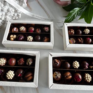 Puede incluir: Cuatro cajas blancas texturizadas llenas de bombones variados. Cada caja contiene dos filas de bombones con varios ingredientes, incluyendo nueces, perlas de chocolate blanco y flores secas. Una cinta con el texto "Petal & Cocoa" es visible.