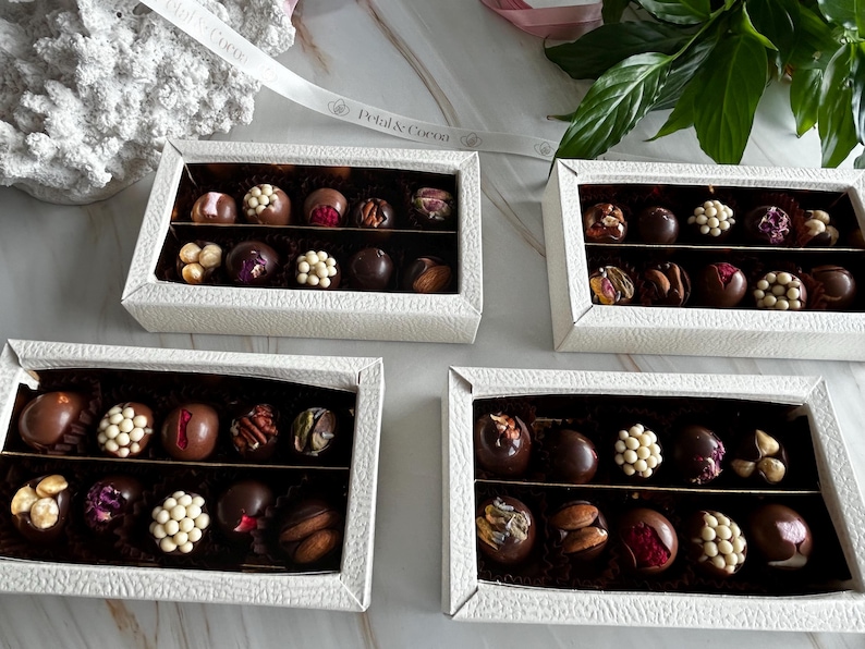 Puede incluir: Cuatro cajas blancas texturizadas llenas de bombones variados. Cada caja contiene dos filas de bombones con varios ingredientes, incluyendo nueces, perlas de chocolate blanco y flores secas. Una cinta con el texto "Petal & Cocoa" es visible.