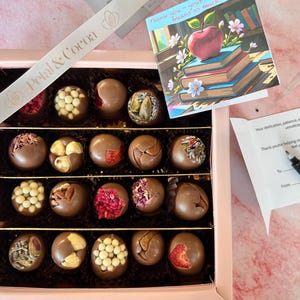 Puede incluir: Una caja de regalo rosa llena de trufas de chocolate variadas, algunas decoradas con nueces, bayas y otros ingredientes. Tambi&eacute;n se ve una tarjeta con una ilustraci&oacute;n de manzana y libros y una cinta con el texto "Petal & Cocoa".