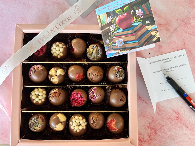 Puede incluir: Una caja de regalo rosa llena de trufas de chocolate variadas, algunas decoradas con nueces, bayas y otros ingredientes. Tambi&eacute;n se ve una tarjeta con una ilustraci&oacute;n de manzana y libros y una cinta con el texto "Petal & Cocoa".